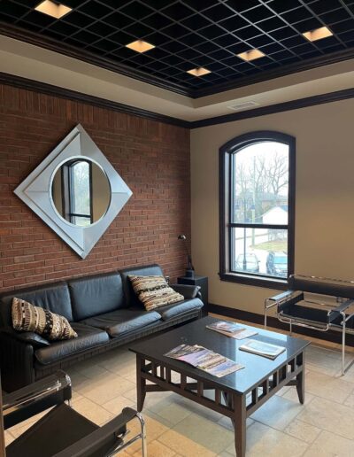 An elegant waiting area with a black sofa, decorative pillows, a modern coffee table, and a unique mirror on a brick wall.