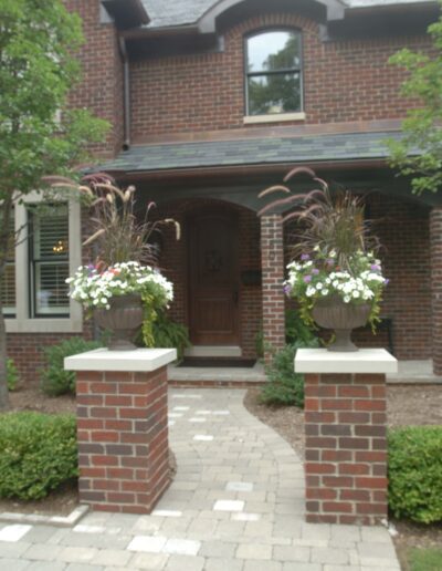 A brick house entrance with potted flowers on pillars flanking a stone pathway.