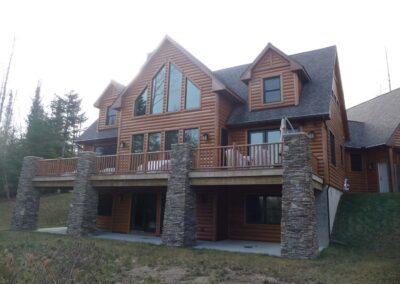 A large two-story wooden house with stone foundation accents and a balcony, surrounded by trees.